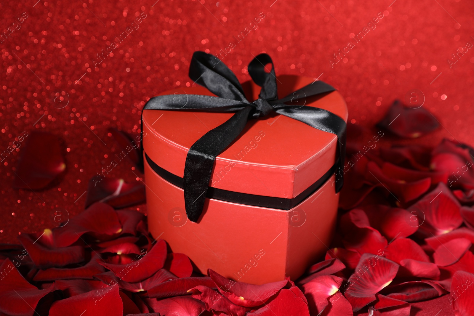 Photo of Beautiful gift box in shape of heart and rose petals on red glitter background, closeup. Happy Valentine's day