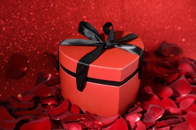 Beautiful gift box in shape of heart and rose petals on red glitter background, closeup. Happy Valentine's day