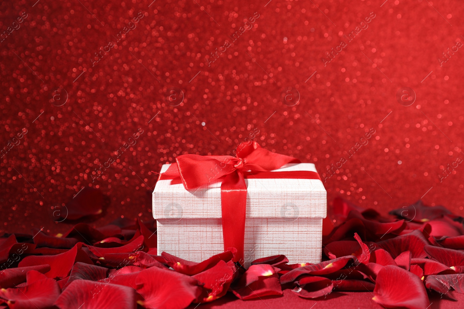 Photo of Beautiful gift box and rose petals on red glitter background, closeup. Romantic present