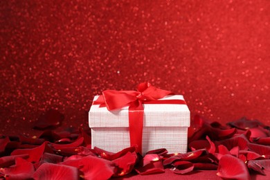 Beautiful gift box and rose petals on red glitter background, closeup. Romantic present