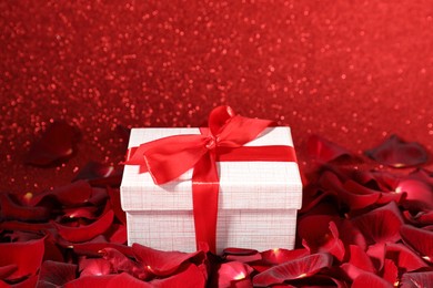 Photo of Beautiful gift box and rose petals on red glitter background, closeup. Romantic present