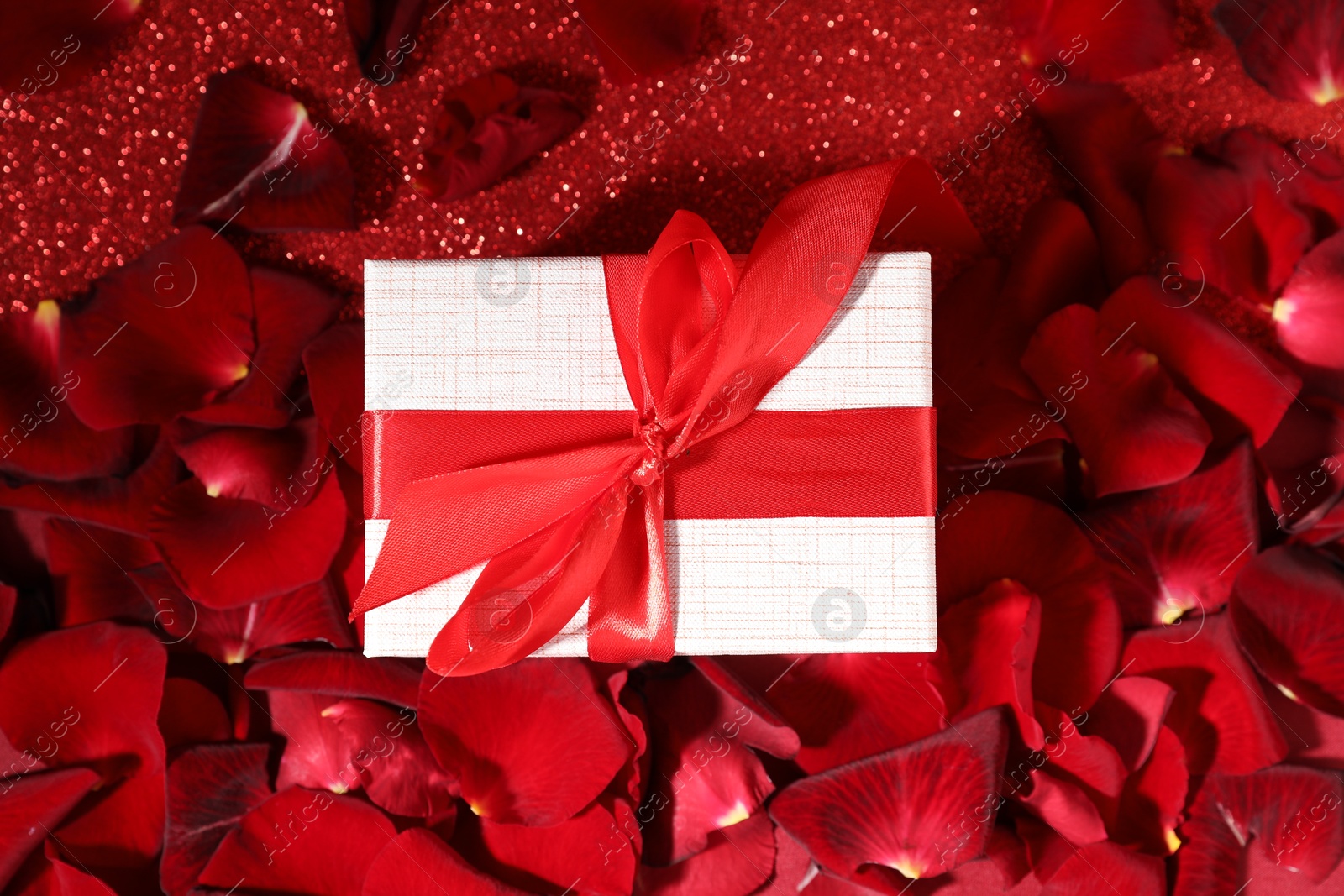 Photo of Beautiful gift box and rose petals on red glitter background, flat lay. Romantic present