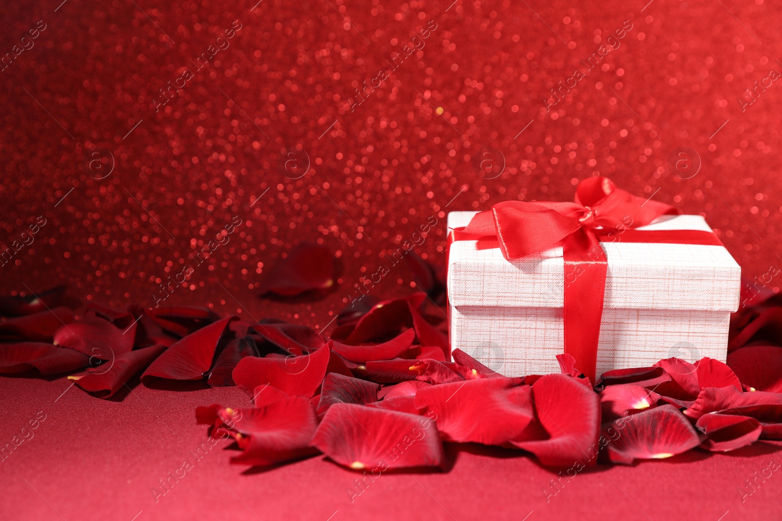 Photo of Beautiful gift box and rose petals on red surface against glitter background, closeup. Romantic present