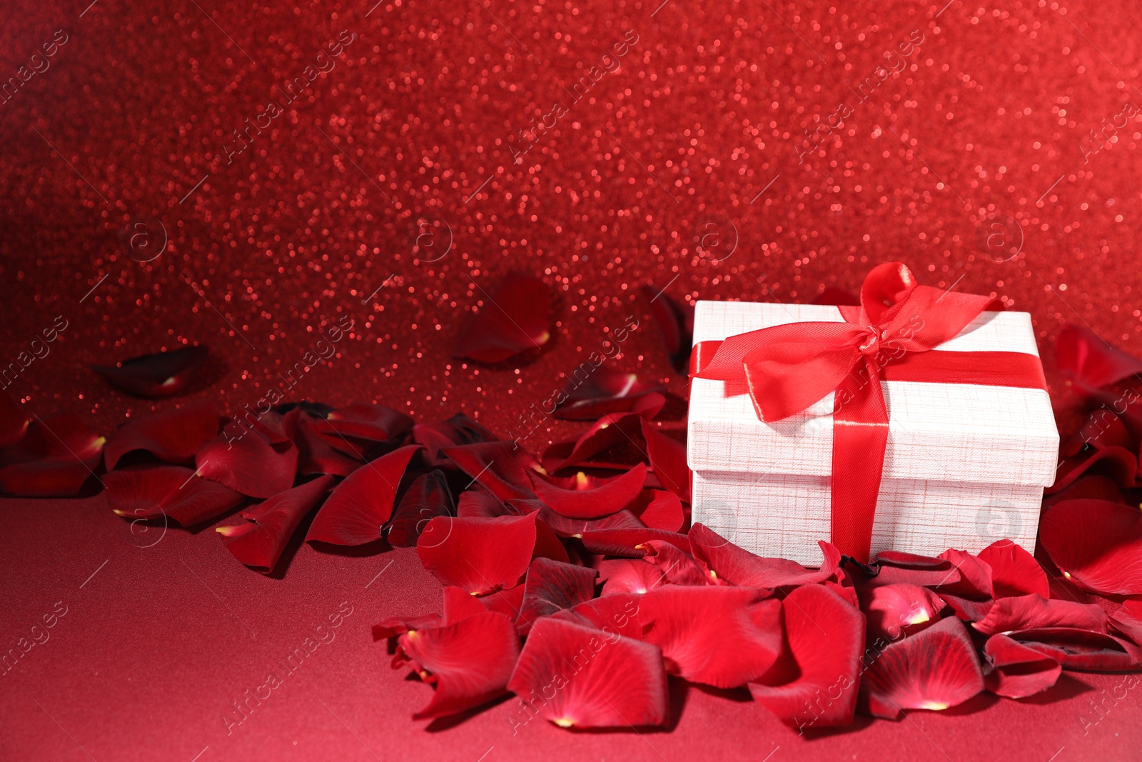 Photo of Beautiful gift box and rose petals on red surface against glitter background. Romantic present