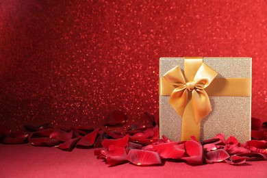 Photo of Beautiful gift box and rose petals on red surface against glitter background, space for text. Romantic present