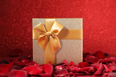 Photo of Beautiful gift box among rose petals on red glitter background, closeup. Romantic present