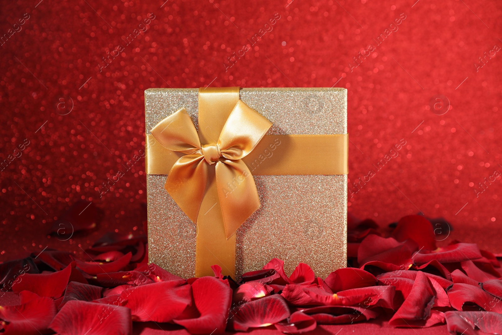 Photo of Beautiful gift box among rose petals on red glitter background, closeup. Romantic present