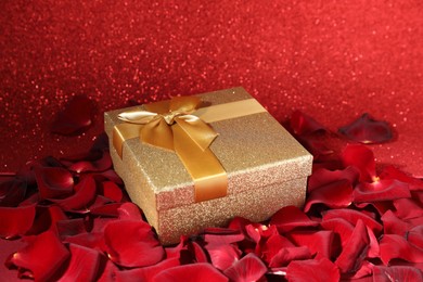 Photo of Beautiful gift box among rose petals on red glitter background, closeup. Romantic present