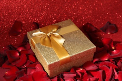 Photo of Beautiful gift box among rose petals on red glitter background, closeup. Romantic present