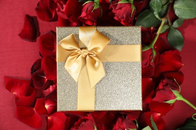 Beautiful gift box among rose flowers on red background, flat lay. Romantic present