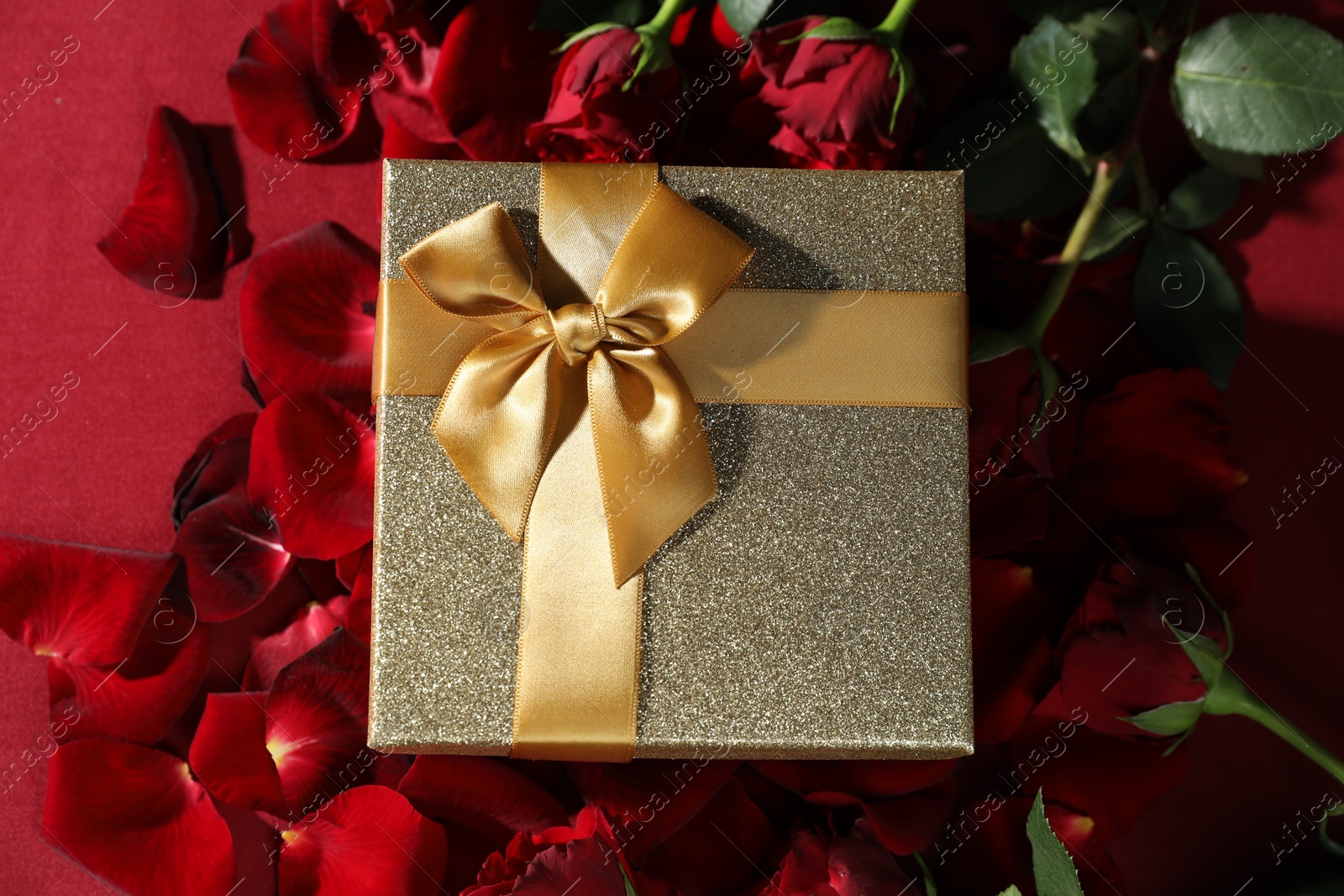 Photo of Beautiful gift box among rose flowers on red background, flat lay. Romantic present