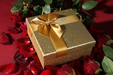 Beautiful gift box among rose flowers and petals on red background, closeup. Romantic present