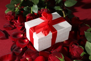 Beautiful gift box among rose flowers on red background, closeup. Romantic present