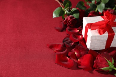 Photo of Beautiful gift box among rose flowers and petals on red background, space for text. Romantic present