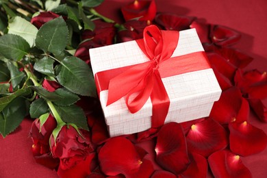 Photo of Beautiful gift box among rose flowers and petals on red background. Romantic present