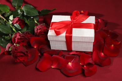 Beautiful gift box among rose flowers and petals on red background. Romantic present