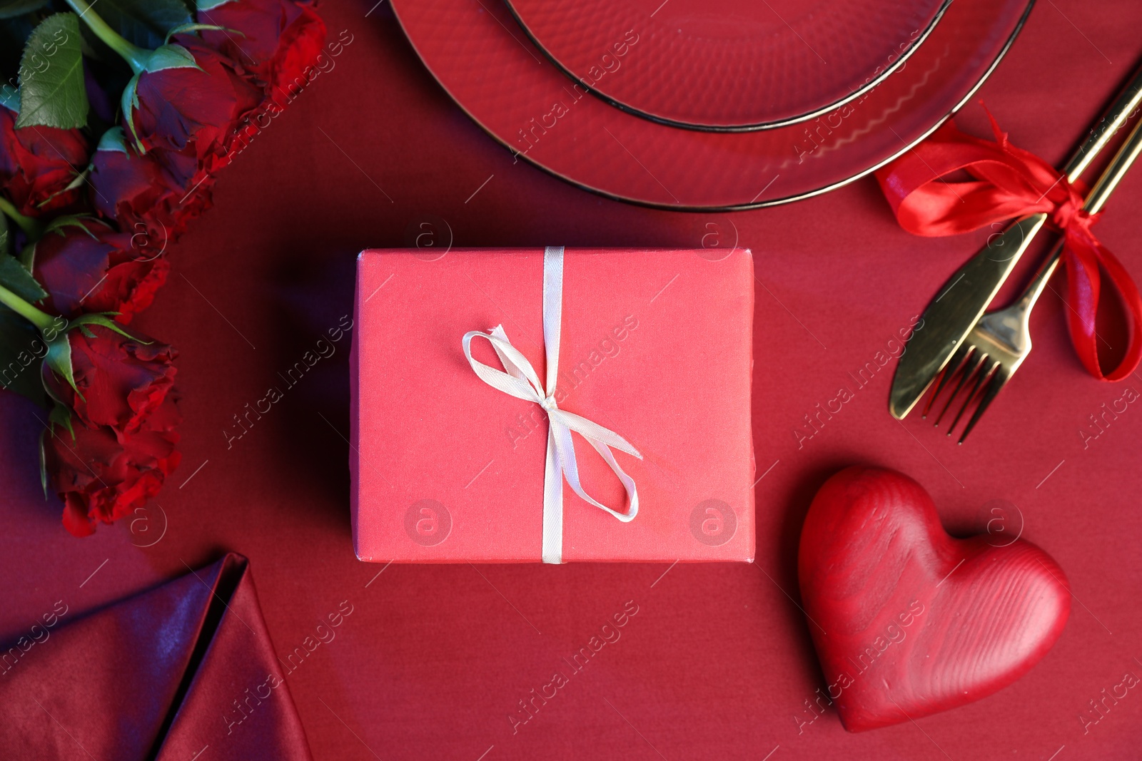 Photo of Gift box, roses and tableware on red table, flat lay. Table setting for romantic Valentine's day celebration