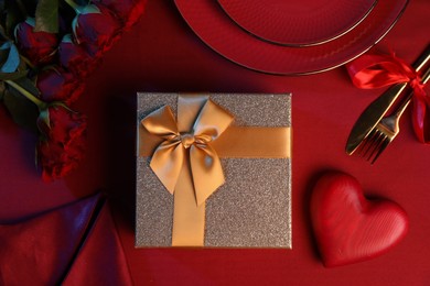 Photo of Gift box, roses and tableware on red table, flat lay. Table setting for romantic Valentine's day celebration