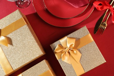 Gift boxes and tableware on red table, flat lay. Table setting for romantic dinner