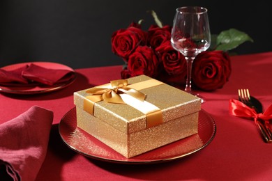 Photo of Gift box, plate and roses on red table, closeup. Table setting for romantic dinner