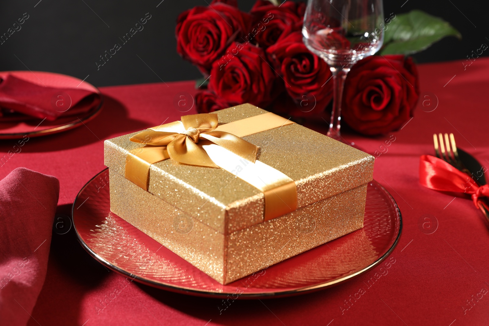 Photo of Gift box, plate and roses on red table, closeup. Table setting for romantic dinner