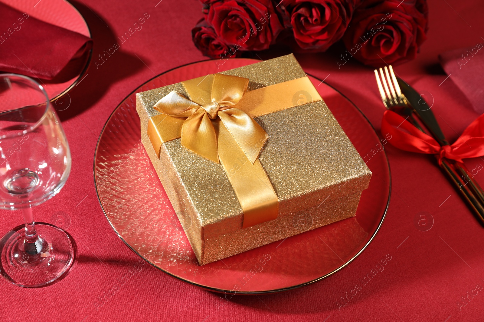 Photo of Gift box, tableware and roses on red table, closeup. Table setting for romantic dinner