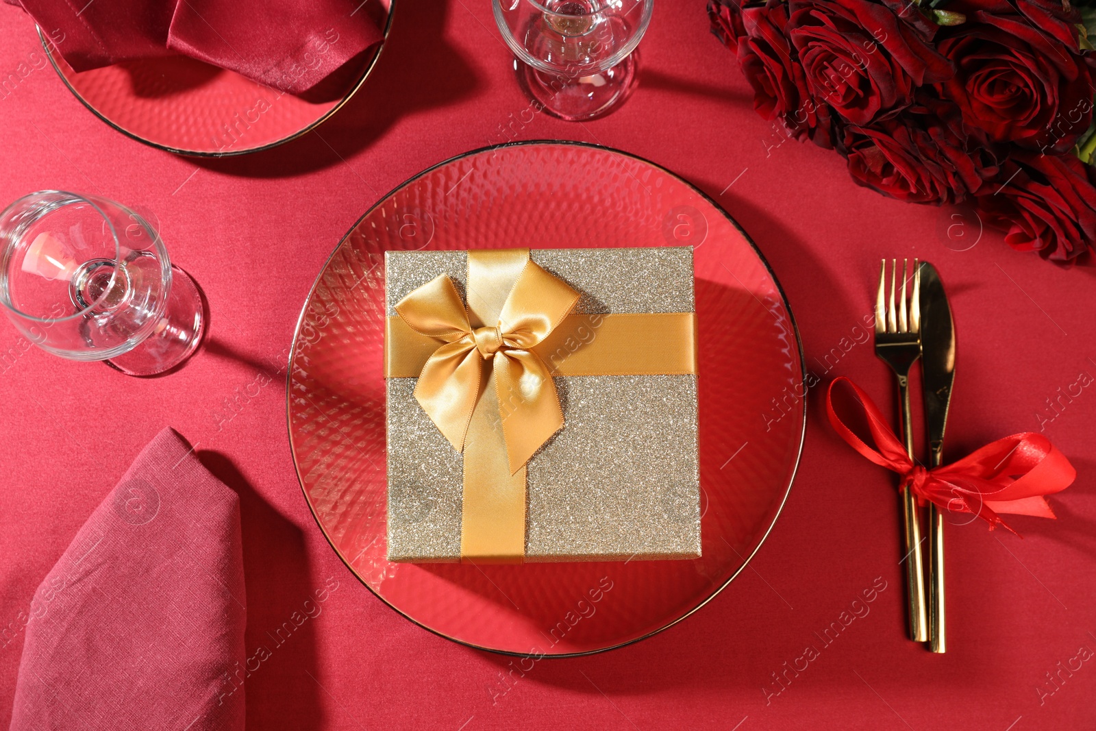 Photo of Gift box, tableware and roses on red table, flat lay. Table setting for romantic dinner