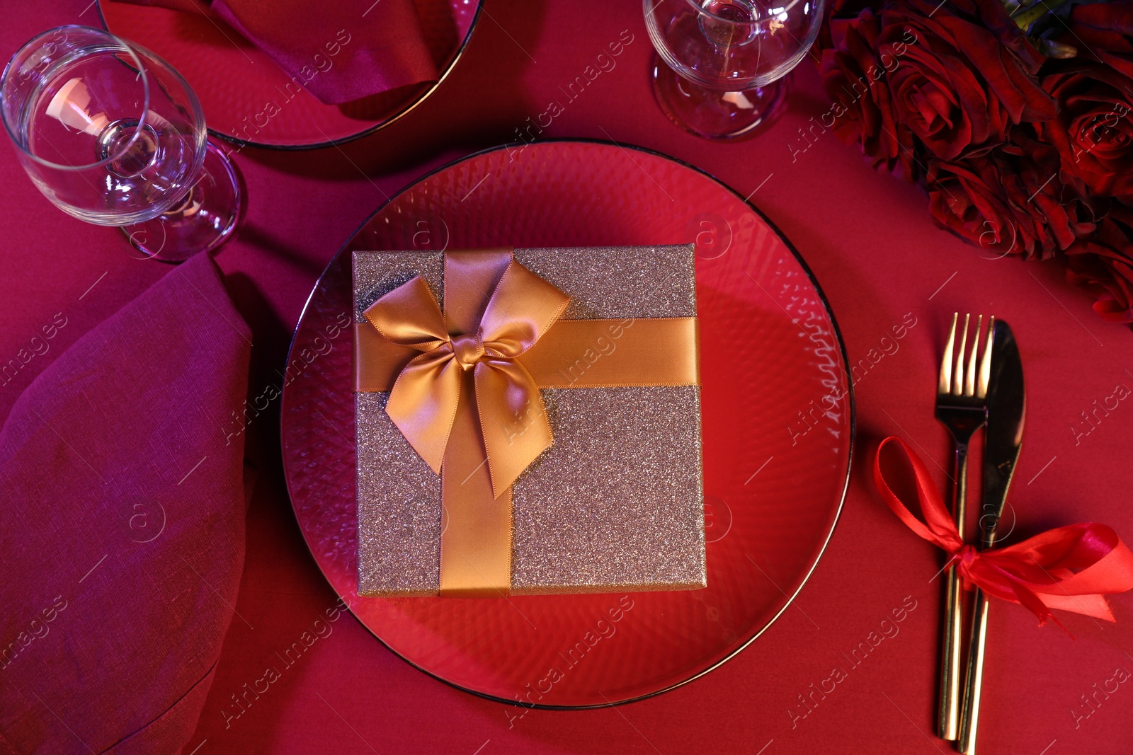 Photo of Gift box, tableware and roses on red table, flat lay. Table setting for romantic dinner