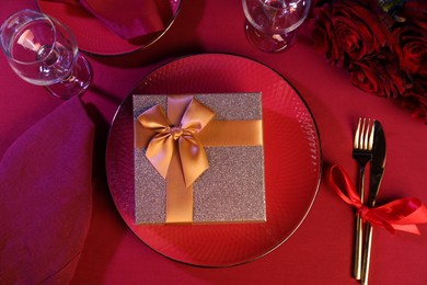 Photo of Gift box, tableware and roses on red table, flat lay. Table setting for romantic dinner