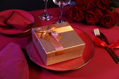 Photo of Gift box, tableware and roses on red table, closeup. Table setting for romantic dinner