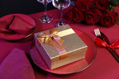 Photo of Gift box, tableware and roses on red table, closeup. Table setting for romantic dinner