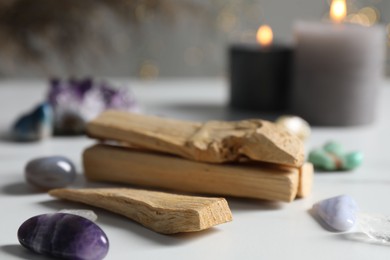 Photo of Palo santo sticks, gemstones and burning candles on white table against blurred lights, closeup