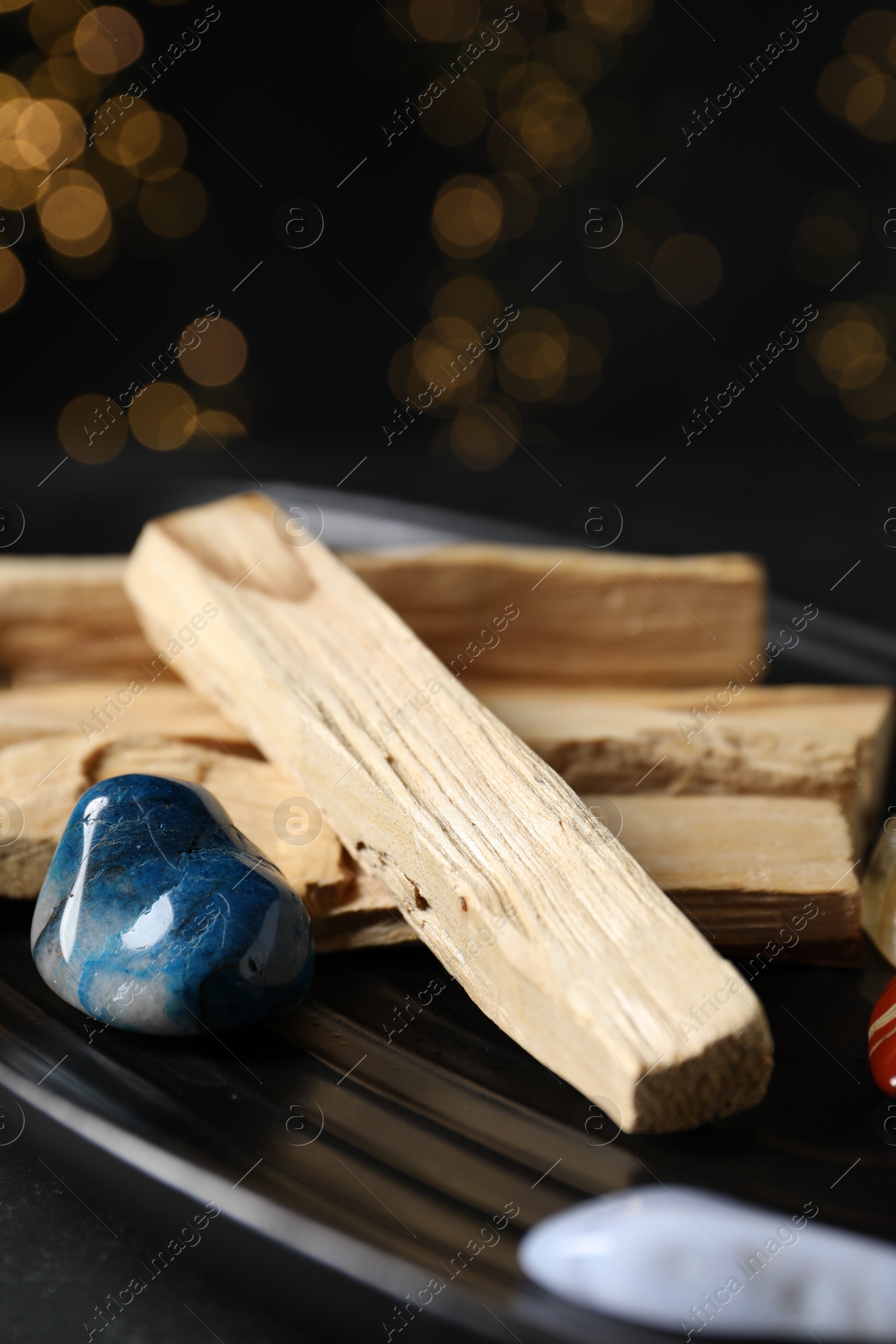 Photo of Palo santo sticks and gemstones on black table against blurred lights, closeup