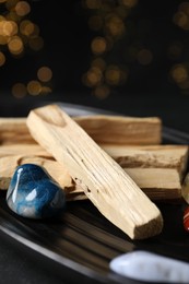 Photo of Palo santo sticks and gemstones on black table against blurred lights, closeup