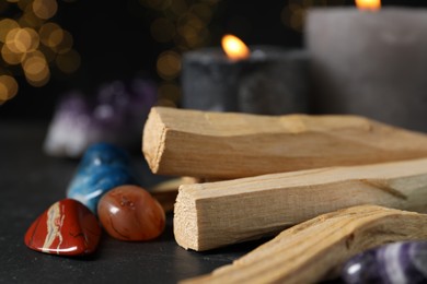 Photo of Palo santo sticks, gemstones and burning candles on black table against blurred lights, closeup