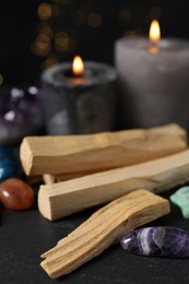 Photo of Palo santo sticks, gemstones and burning candles on black table against blurred lights, closeup