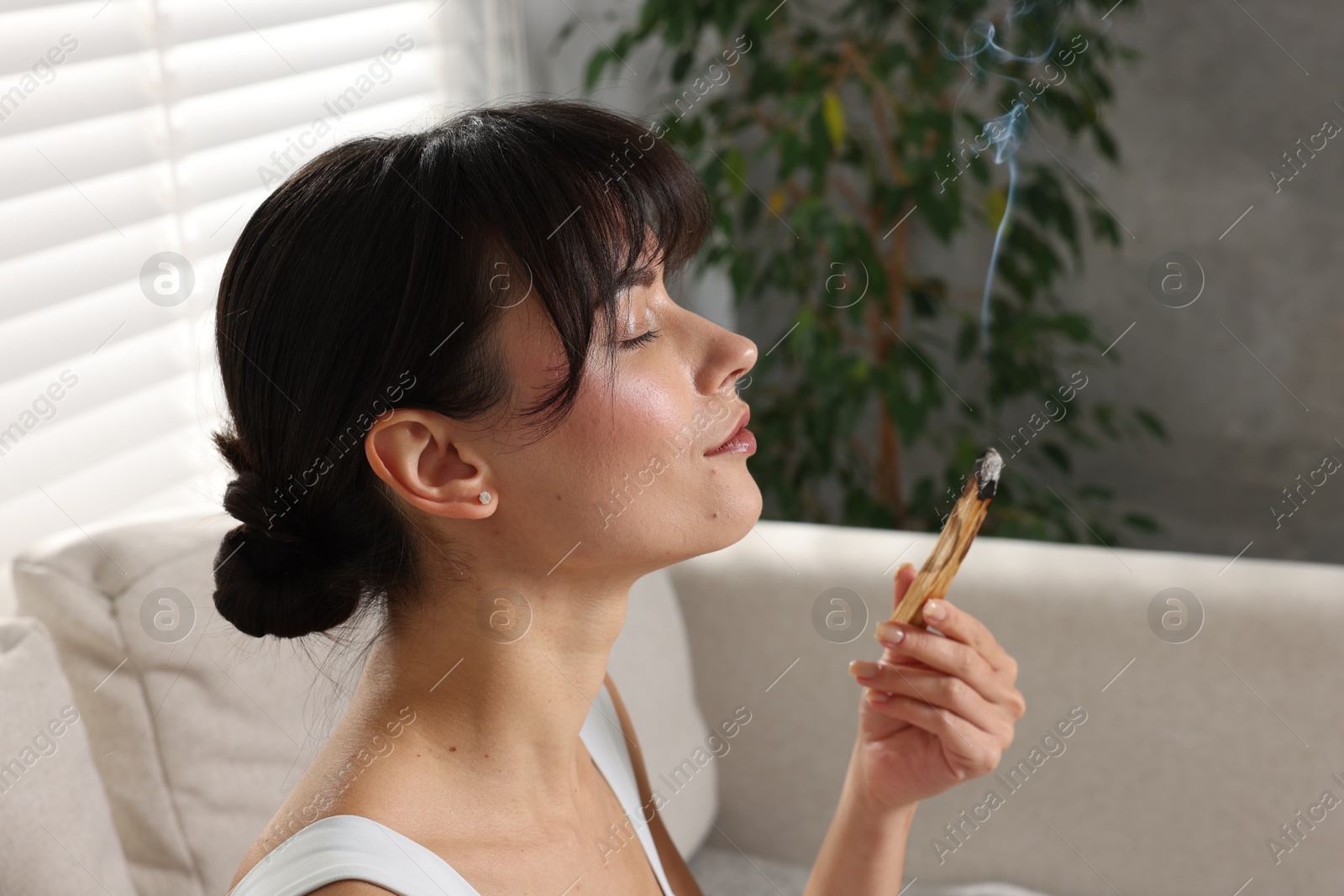 Photo of Woman with smoldering palo santo stick on sofa at home