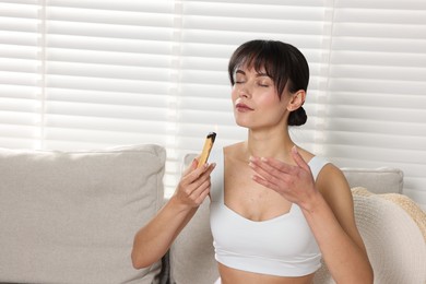 Photo of Woman with burning palo santo stick on sofa at home, space for text