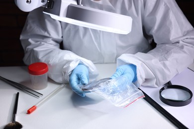 Photo of Forensic expert examining evidence in plastic bag at table indoors, closeup