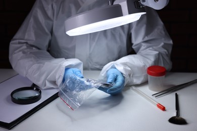 Photo of Forensic expert examining evidence in plastic bag at table indoors, closeup