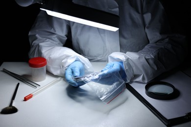 Photo of Forensic expert examining evidence in plastic bag at table indoors, closeup