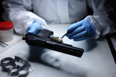 Photo of Forensic expert taking fingerprints with brush from gun at table indoors, closeup