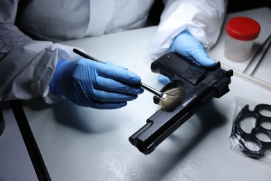 Photo of Forensic expert taking fingerprints with brush from gun at table indoors, closeup