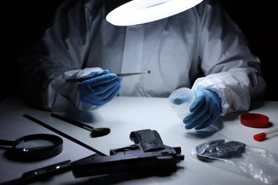 Photo of Forensic expert holding tweezers with hair sample and plastic container at table indoors