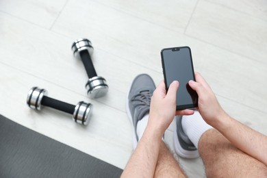 Photo of Man with smartphone and dumbbells on floor indoors, above view