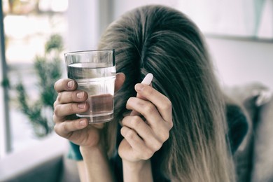 Sad young woman with pill and glass of water indoors, closeup. Toned for dramatic atmosphere