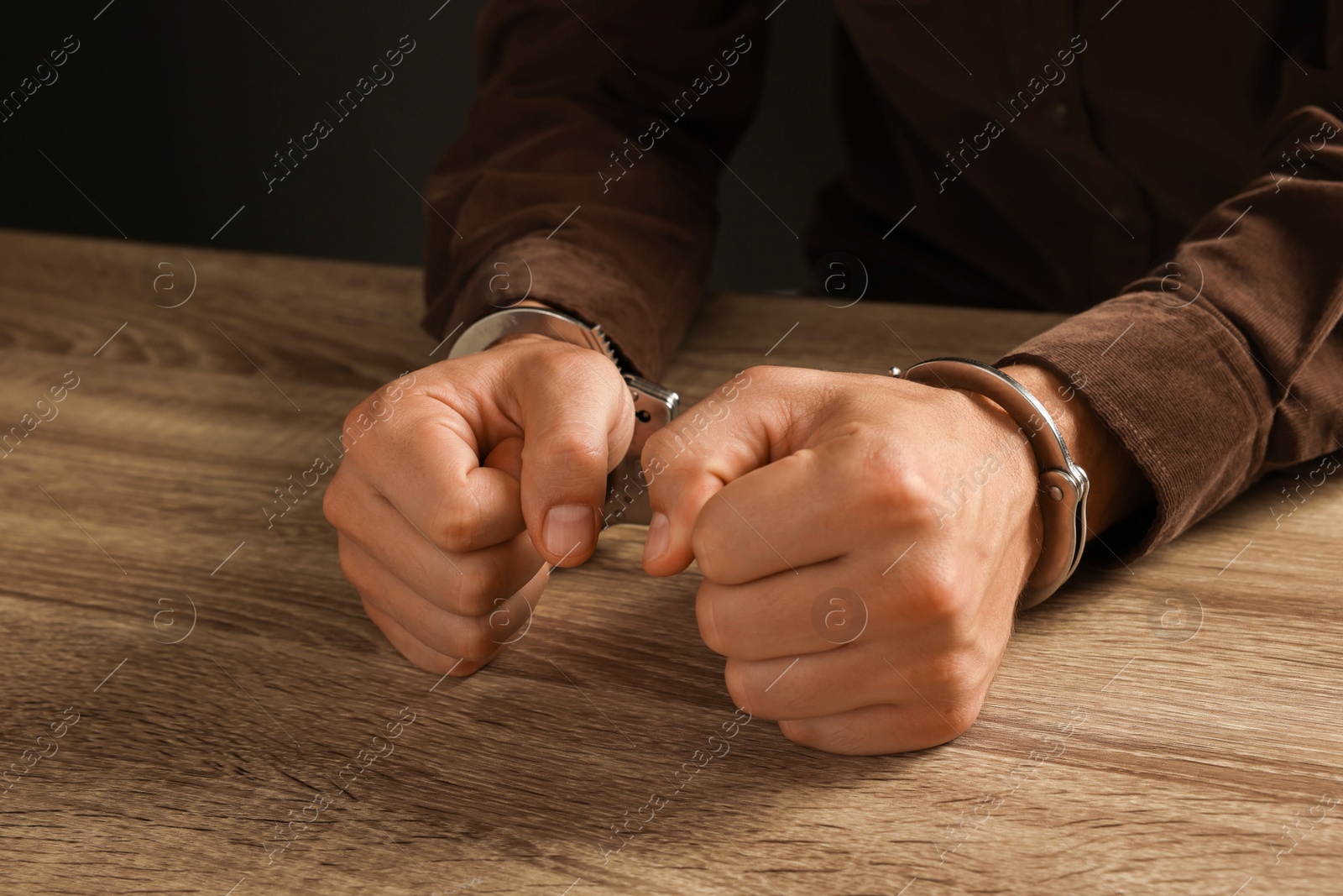 Photo of Man in metal handcuffs at wooden table against black background, closeup