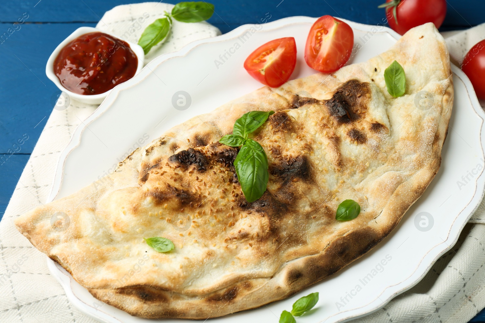 Photo of Tasty calzone with basil, tomatoes and sauce on blue wooden table, flat lay