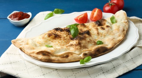 Photo of Tasty calzone with basil, tomatoes and sauce on blue wooden table, closeup