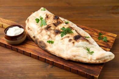 Photo of Board with tasty calzone, parsley and sauce on wooden table, closeup